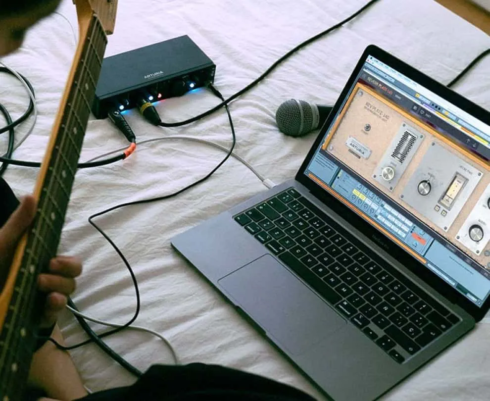 Set de ordenador portátil mac, guitarra e interfaz de audio Arturia Minifuse 4 sobre una cama con sábana blanca