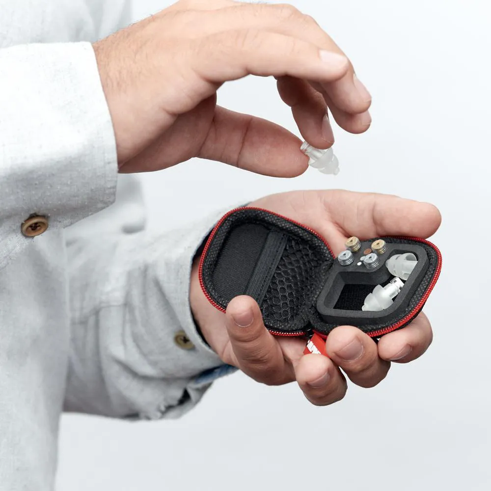 Manos sosteniendo un tapón auditivo transparente Alpine MusicSafe Pro a punto de guardarlo en su estuche compacto con compartimentos de filtros y malla organizadora.